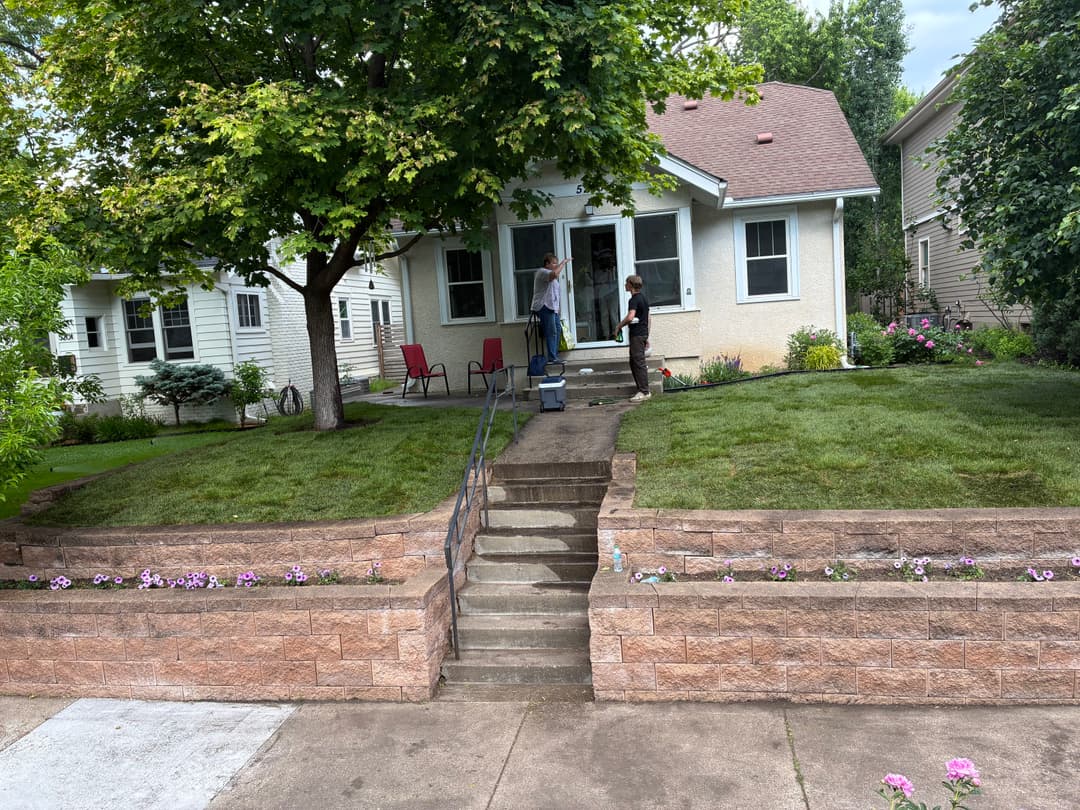 Home exterior with well-maintained lawn, flower beds, and two people on the porch steps.