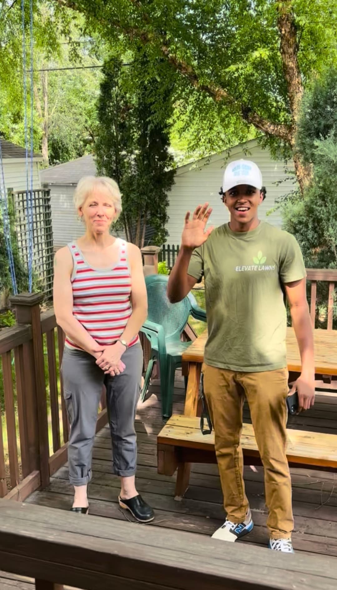 Woman and young man on a deck, both smiling and waving, in a backyard setting.