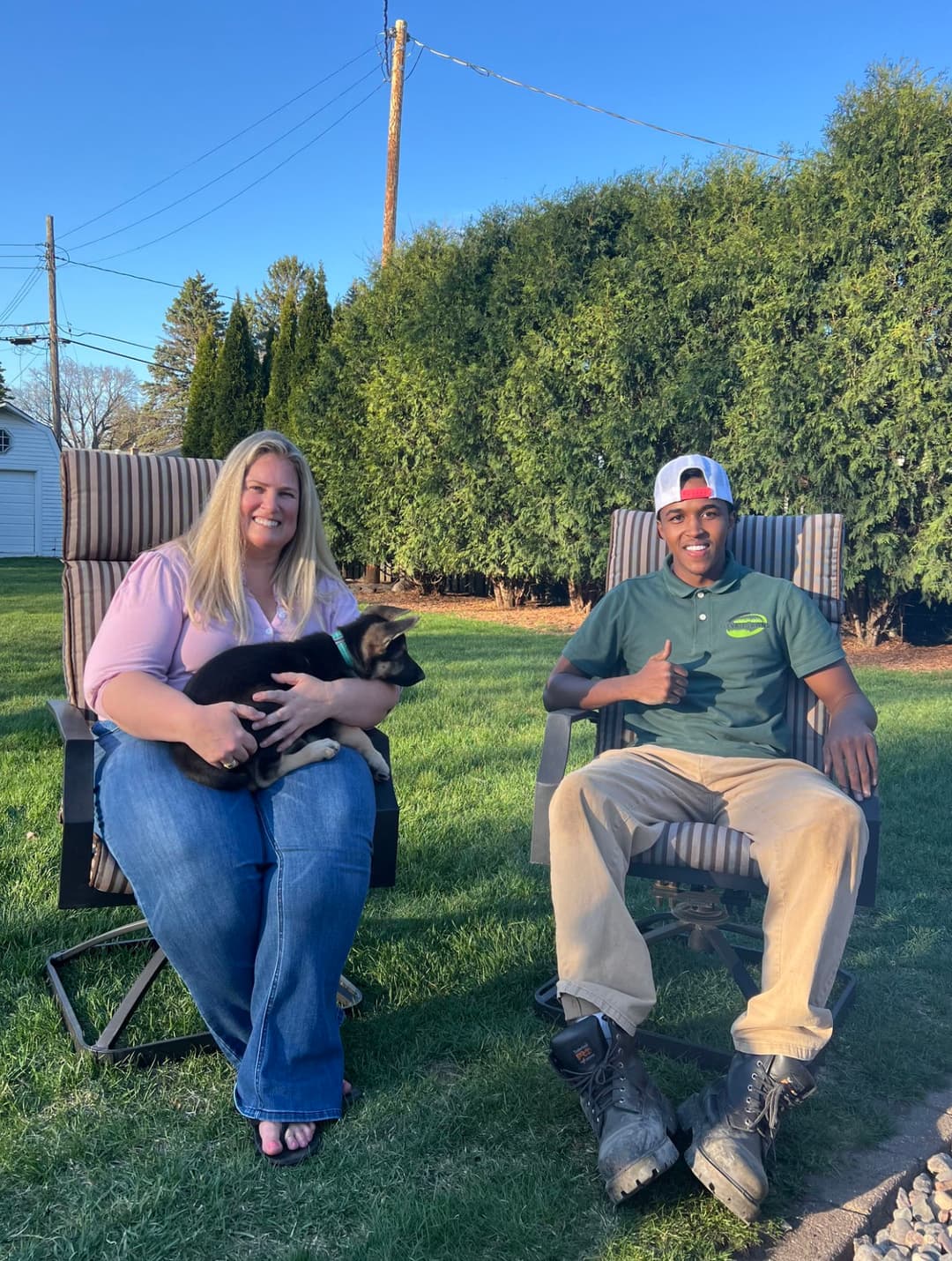 Woman holding puppy and man giving thumbs up sit in lawn chairs outdoors.