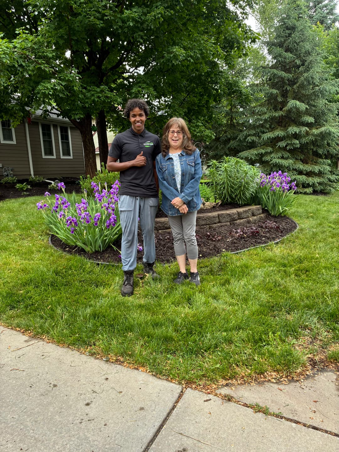 Two people standing in a landscaped garden with purple flowers and green shrubs.