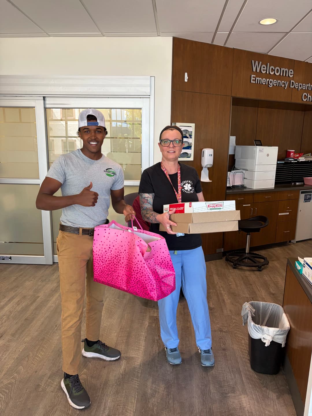 Man and woman in hospital lobby holding gifts and food items, smiling during a delivery.