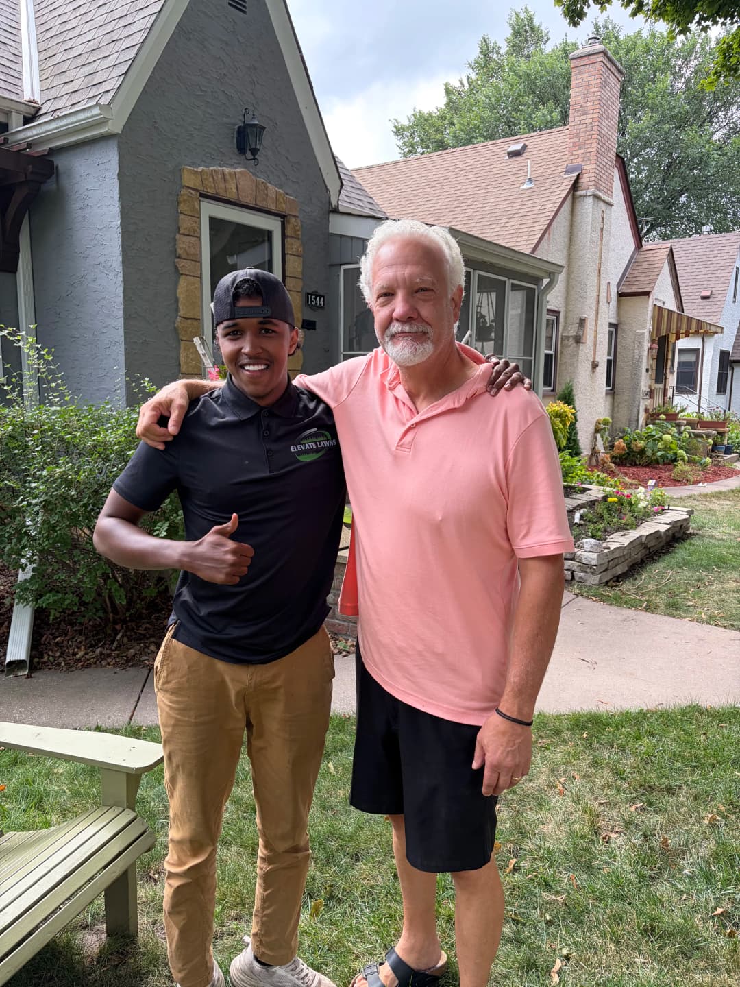 Two men smiling outdoors in front of a house, one giving a thumbs up, showcasing a friendly interaction.
