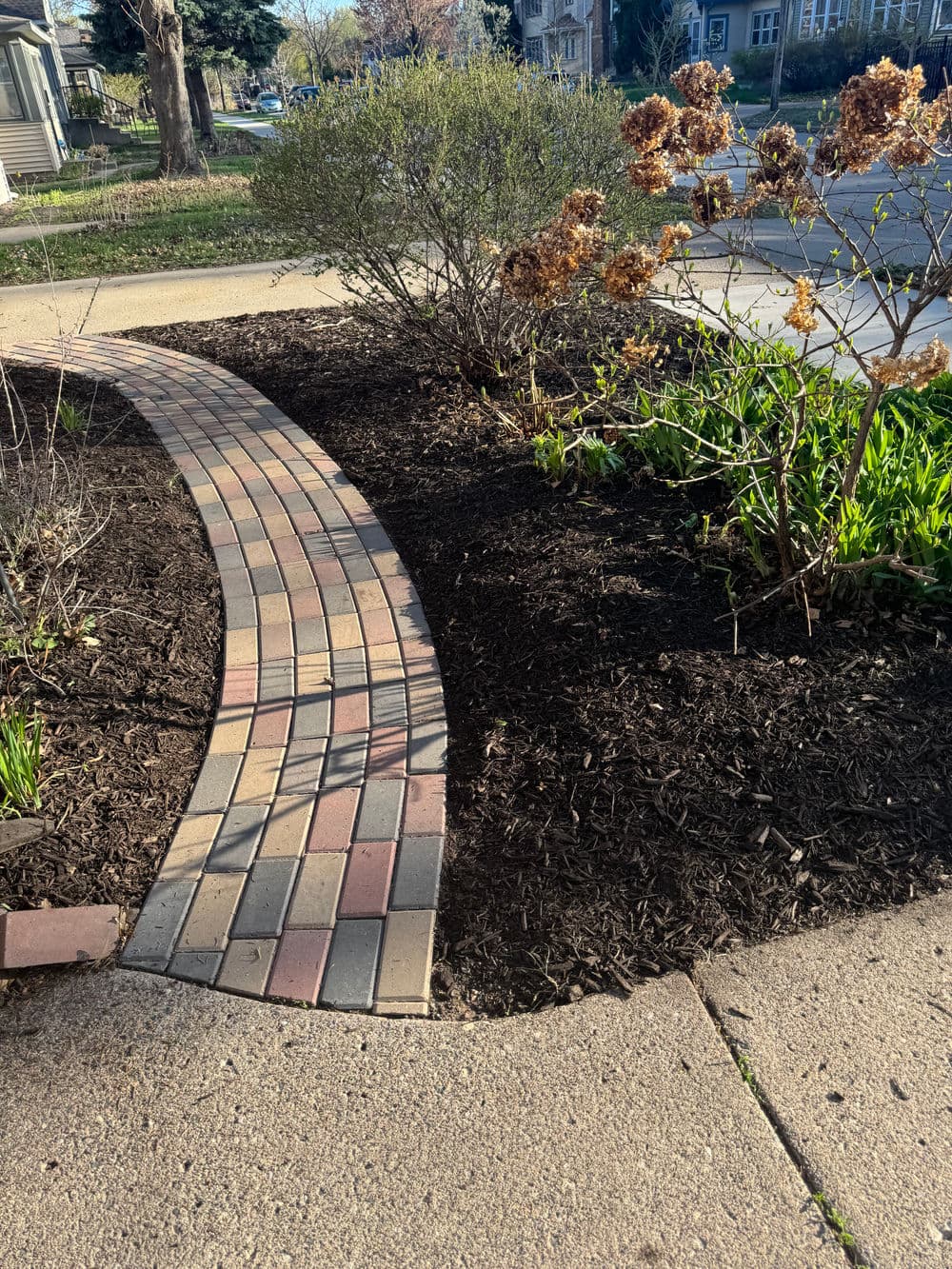 Curved brick walkway through landscaped garden with mulch and flowering plants.