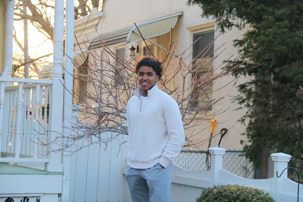 Young man in a white sweater standing outside near a bare tree at sunset.