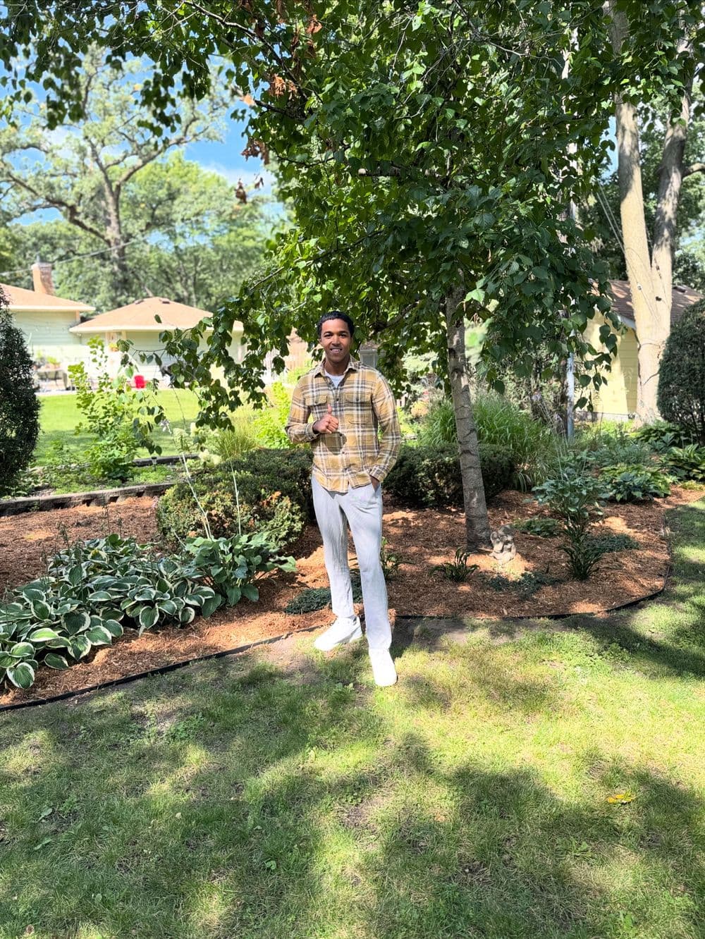 Man in plaid shirt stands in a vibrant garden with lush greenery and flowers.